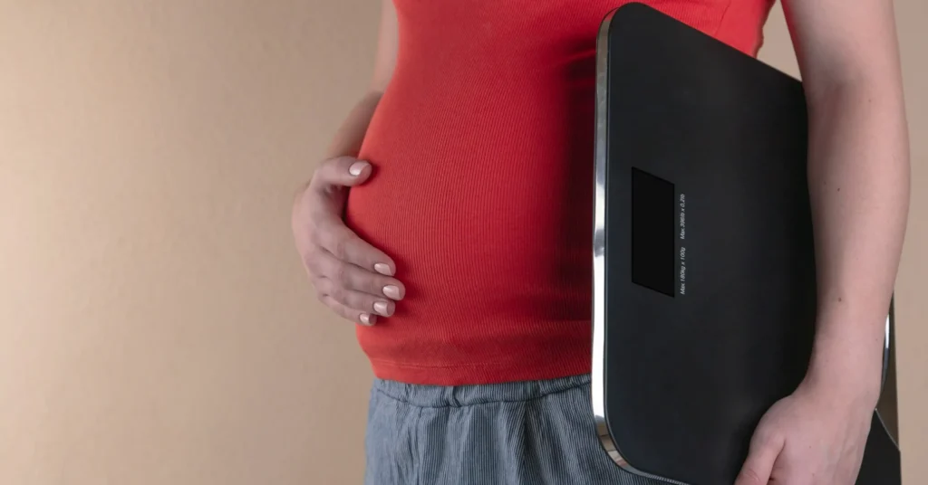 A person holding a weighing balance in one hand and put another hand on the belly.